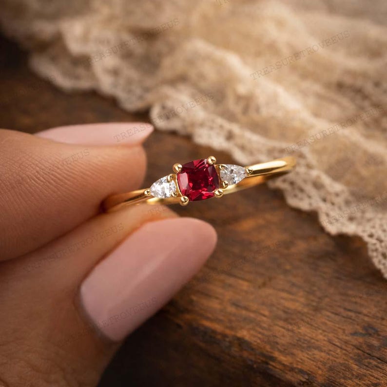 Puede incluir: Un anillo de oro con una gema roja cuadrada en el centro, flanqueada por dos piedras blancas triangulares m&aacute;s peque&ntilde;as. El dise&ntilde;o del anillo se destaca sobre un fondo borroso, resaltando los colores.