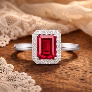 以下が含まれることがあります： 長方形の鮮やかな赤い宝石をあしらったシルバーリング。小さな透明な石のハローで囲まれています。リングは木製の表面に置かれ、ジュエリーのエレガントなデザインを際立たせています。