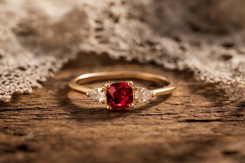 Puede incluir: Un anillo de oro con una piedra central roja oscura de talla coj&iacute;n y dos piedras blancas m&aacute;s peque&ntilde;as en forma de pera a cada lado. El anillo est&aacute; sobre una superficie de madera con una tela de encaje en el fondo.