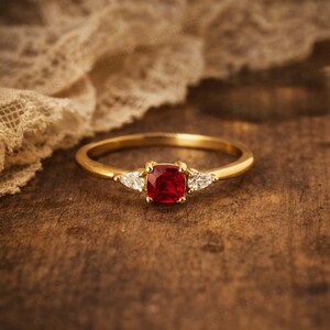 Puede incluir: Un anillo de oro con una gema roja cuadrada en el centro y dos gemas blancas triangulares m&aacute;s peque&ntilde;as a cada lado. El anillo est&aacute; sobre una superficie de madera oscura.