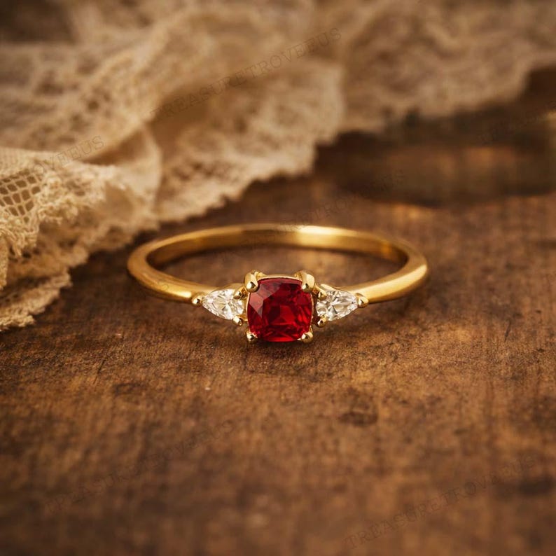 Puede incluir: Un anillo de oro con una gema roja cuadrada en el centro y dos gemas blancas triangulares m&aacute;s peque&ntilde;as a cada lado. El anillo est&aacute; sobre una superficie de madera oscura.