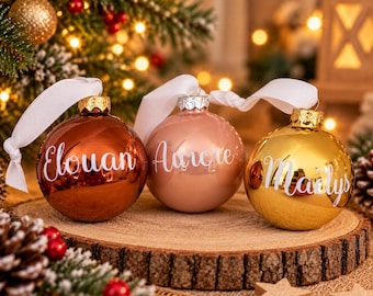 Könnte beinhalten: Drei kugelförmige Weihnachtskugeln mit weißen Bändern. Die linke Kugel ist kupferfarben mit dem Namen Elouan, die mittlere ist roségold mit dem Namen Aurore und die rechte ist goldfarben mit dem Namen Maëlys in weißer Schreibschrift.