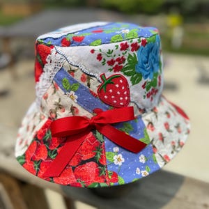May include: A quilted bucket hat featuring a patchwork design of various strawberry-themed fabrics in red, white, blue, and green. The hat is accented with a white lace trim and a prominent red ribbon bow tied at the front center.