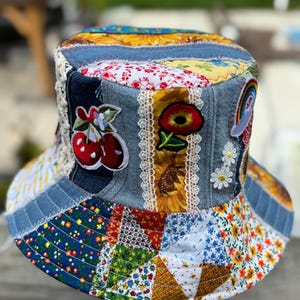 May include: A patchwork bucket hat constructed from various fabric scraps including blue denim, floral prints, and sunflower patterns. The hat features embroidered patches of red cherries, a red poppy, and a rainbow, accented with white lace trim.