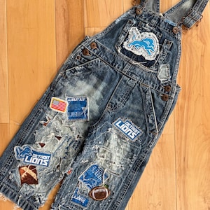 May include: Distressed denim dungarees with patches displaying the Detroit Lions logo, an American flag, and a football. The blue denim overalls have ripped and frayed details, with adjustable straps and metal hardware.