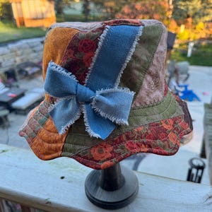 May include: A patchwork bucket hat featuring various fabric panels in shades of orange, olive green, and floral patterns. The hat is accented with a prominent blue denim bow with frayed edges, stitched onto the front of the quilted crown.