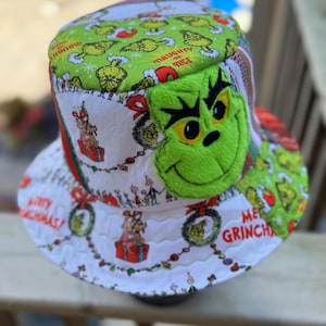 May include: A quilted bucket hat featuring a green and white fabric pattern with illustrations of the Grinch, Christmas wreaths, and text reading Naughty or Nice and Merry Grinchmas. A large, plush, green embroidered Grinch face is attached to the side.