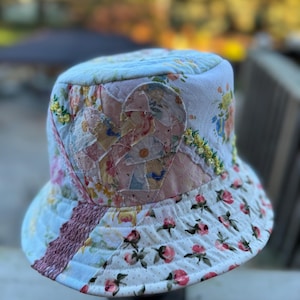 May include: A quilted bucket hat featuring a patchwork design of various floral fabrics in shades of light blue, pink, and white. The hat includes a heart-shaped fabric appliqué, decorative yellow embroidery, and a textured brown fabric strip.