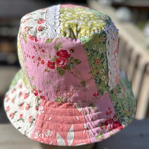 May include: A patchwork bucket hat constructed from various floral-patterned fabric panels in shades of pink, green, and white. The hat features vertical strips of white lace trim between the fabric sections and visible quilted stitching on the brim.