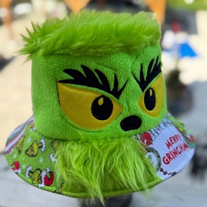May include: A bright green fuzzy bucket hat featuring an embroidered face with yellow eyes and black eyebrows. The brim displays a repeating pattern of green characters in red hats and a white patch with the red text Merry Grinchmas.