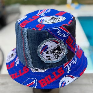 Op de afbeelding: Handgemaakte vissershoed met een patchwork-ontwerp. De hoed combineert denim en blauwe stof bedrukt met het woord "BILLS" in rood en een logo van een footballhelm. De rand en bovenkant zijn blauw met het Bills-logo.