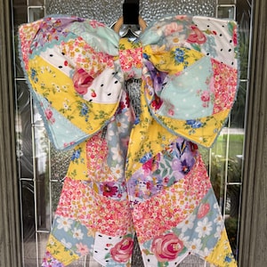 May include: A large decorative bow constructed from various fabric patches featuring floral, polka dot, and strawberry patterns in yellow, pink, blue, and white. The bow has two long hanging tails and is attached to a circular wooden ring for hanging.