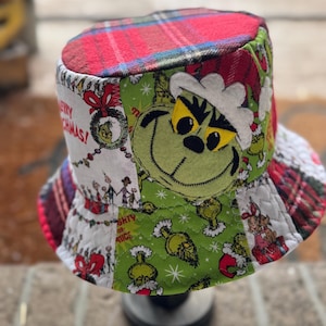 May include: A bucket hat featuring a patchwork design with a red and blue plaid top. The sides display various green Grinch-themed fabric panels, including a large Grinch face, Christmas imagery, and text that reads Merry Christmas.