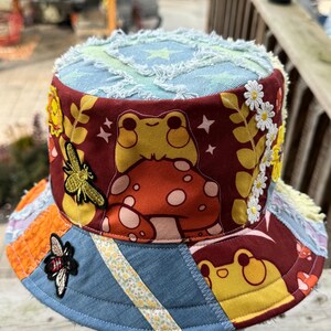 May include: A patchwork bucket hat featuring a dark red fabric with a printed yellow frog sitting on a mushroom, accented by embroidered bee patches, white daisy trim, and blue denim panels with frayed edges and star patterns.
