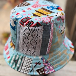 Può includere: Un cappello a secchiello in tessuto azzurro con una stampa multicolore di architetture di ispirazione giapponese, fiori di ciliegio e motivi geometrici, accentuato da una striscia verticale di pizzo bianco all'uncinetto e tessuto grigio.