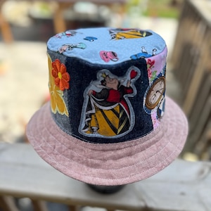 May include: A bucket hat featuring a light blue top panel with a multi-colored character print. The side panels are made of dark denim fabric with embroidered patches of a queen, a clock, and a flower. The brim is a solid dusty pink fabric.