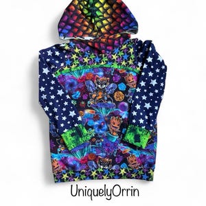 May include: A long-sleeved hoodie with a colorful design. The hood features a rainbow scale pattern. The body of the hoodie has a vibrant print with cartoon characters, stars, and other graphics. The sleeves are navy blue with white stars. The text "UniquelyOrrin" is at the bottom.