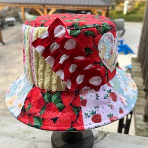 May include: A patchwork bucket hat featuring a mix of fabrics including red strawberry prints, yellow textured fabric, light blue floral patterns, and a red and white polka dot ruffled accent piece attached to the side of the crown.