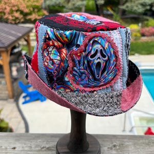 May include: A patchwork bucket hat featuring a vibrant, multi-colored graphic print of horror movie characters, including a masked figure making a peace sign. The hat is constructed from various fabric panels in red, black, and denim, with visible stitching.