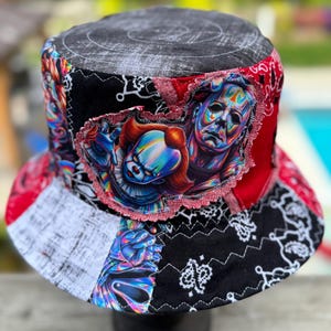 May include: A patchwork bucket hat featuring a black and white distressed top and a colorful graphic print of Pennywise and Michael Myers. The hat is constructed from various fabric panels including red and black paisley patterns and zigzag stitching.