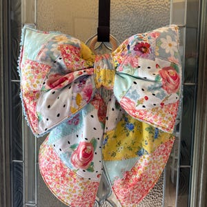 May include: A large, decorative bow made from a patchwork of various floral and polka-dot patterned fabrics in shades of pink, yellow, blue, and white. The bow is attached to a small wooden ring and hangs from a black strap against a glass door.