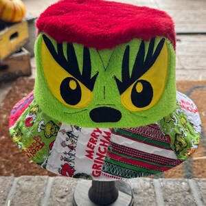 May include: A bucket hat featuring a bright red faux fur top and a lime green plush face with large yellow eyes and black pupils. The brim is made of quilted fabric patches with green, white, and red patterns, including the text Merry Grinchmas.