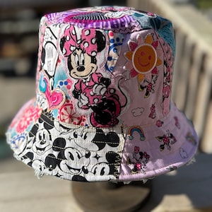 May include: A bucket hat featuring a patchwork design with various Disney character prints. The fabric includes images of Minnie Mouse in pink, a smiling sun, and a black and white pattern of Mickey Mouse faces, accented with hearts and rainbows.