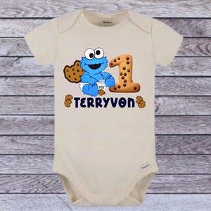 Pu&ograve; includere: Un body da bambino beige con un disegno blu di Cookie Monster che tiene in mano un cartone di latte e un biscotto. Un grande numero 1 fatto di biscotti &egrave; accanto al personaggio. Il nome "TERRYVON" &egrave; stampato in blu navy.