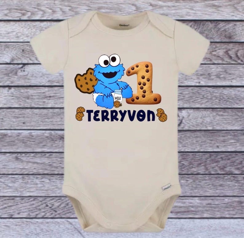 Pu&ograve; includere: Un body da bambino beige con un disegno blu di Cookie Monster che tiene in mano un cartone di latte e un biscotto. Un grande numero 1 fatto di biscotti &egrave; accanto al personaggio. Il nome "TERRYVON" &egrave; stampato in blu navy.