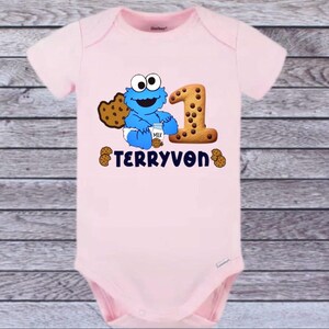 Pu&ograve; includere: Un body rosa chiaro per neonati con un disegno di Cookie Monster blu, un biscotto con gocce di cioccolato e il numero 1 fatto di biscotti. Il nome "TERRYVON" &egrave; stampato sotto.