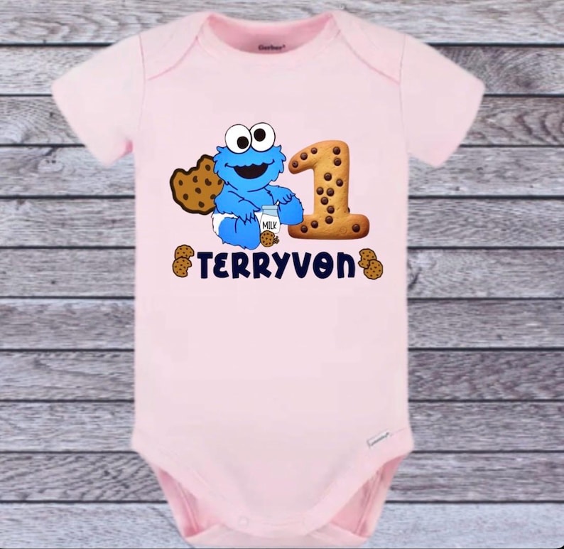 Pu&ograve; includere: Un body rosa chiaro per neonati con un disegno di Cookie Monster blu, un biscotto con gocce di cioccolato e il numero 1 fatto di biscotti. Il nome "TERRYVON" &egrave; stampato sotto.