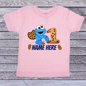 Pu&ograve; includere: T-shirt rosa chiaro con un personaggio dei cartoni animati blu che tiene in mano una bottiglia di latte e un biscotto. Un grande numero 1 fatto di biscotti &egrave; accanto al personaggio. Il testo "NAME HERE" &egrave; stampato sotto.