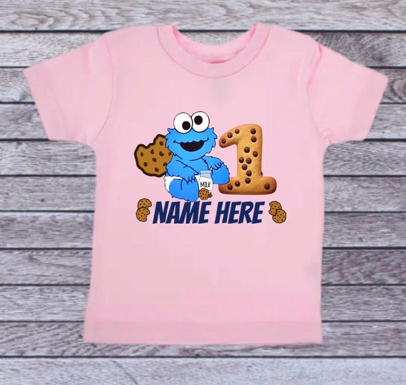 Pu&ograve; includere: T-shirt rosa chiaro con un personaggio dei cartoni animati blu che tiene in mano una bottiglia di latte e un biscotto. Un grande numero 1 fatto di biscotti &egrave; accanto al personaggio. Il testo "NAME HERE" &egrave; stampato sotto.