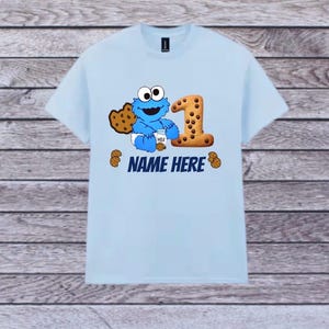 Pu&ograve; includere: T-shirt azzurra con un design di Cookie Monster e il numero 1 fatto di biscotto. Il design include il testo "NAME HERE" e grafiche di biscotti. La maglietta &egrave; a girocollo e maniche corte.