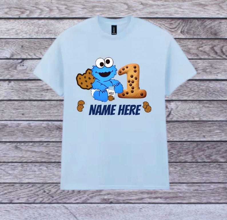 Pu&ograve; includere: T-shirt azzurra con un design di Cookie Monster e il numero 1 fatto di biscotto. Il design include il testo "NAME HERE" e grafiche di biscotti. La maglietta &egrave; a girocollo e maniche corte.