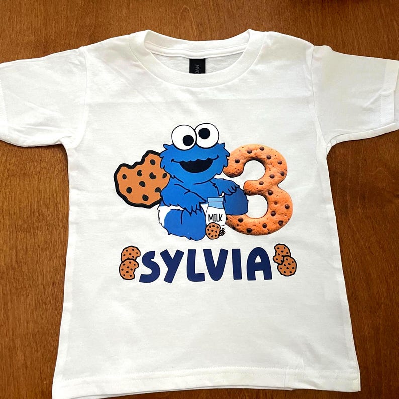 Pu&ograve; includere: Maglietta bianca con un personaggio dei cartoni animati blu, una bottiglia di latte e biscotti. Ha un grande numero 3 e il nome "SYLVIA" in blu. Il numero 3 e i biscotti hanno un design a biscotto marrone.