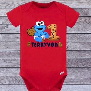 Pu&ograve; includere: Un body rosso per neonati con un motivo di Cookie Monster, un biscotto con il numero 1 e il nome "TERRYVON". Il design include biscotti e un cartone di latte. Un outfit divertente per il primo compleanno.