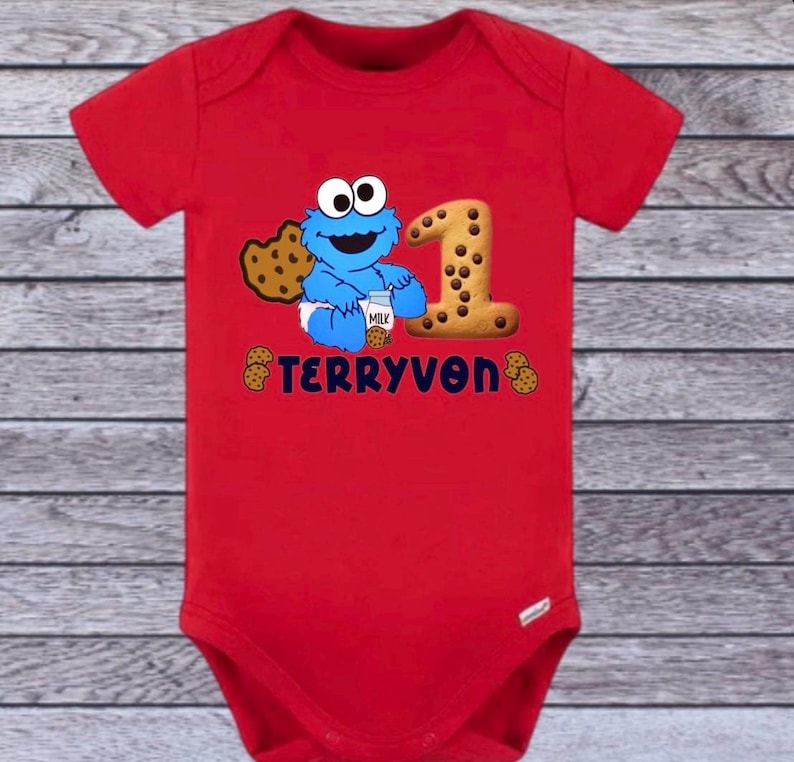 Pu&ograve; includere: Un body rosso per neonati con un motivo di Cookie Monster, un biscotto con il numero 1 e il nome "TERRYVON". Il design include biscotti e un cartone di latte. Un outfit divertente per il primo compleanno.
