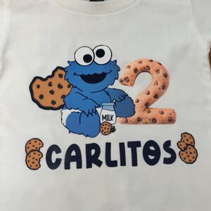 Pu&ograve; includere: T-shirt bianca con un personaggio dei cartoni animati blu, un biscotto e latte. Il numero "2" &egrave; disegnato con un motivo a biscotto. Il nome "CARLITOS" &egrave; stampato sotto, con grafiche di biscotti su entrambi i lati.