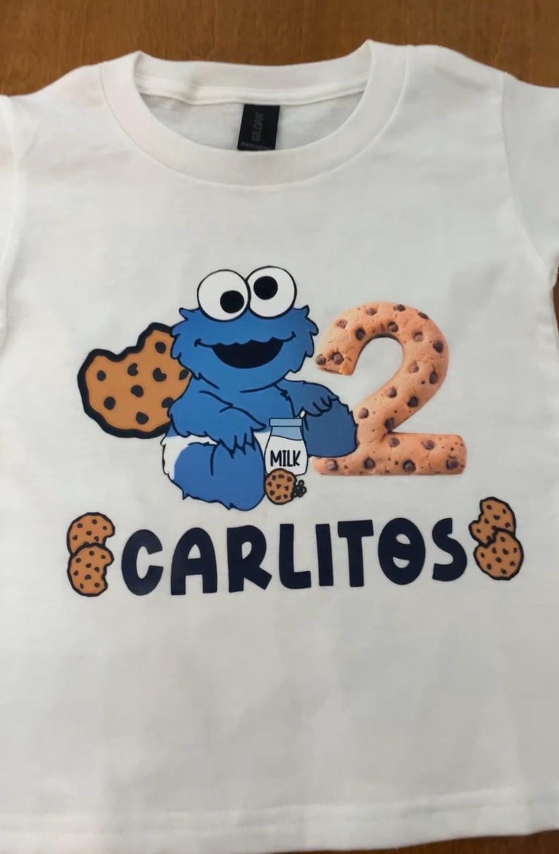 Pu&ograve; includere: T-shirt bianca con un personaggio dei cartoni animati blu, un biscotto e latte. Il numero "2" &egrave; disegnato con un motivo a biscotto. Il nome "CARLITOS" &egrave; stampato sotto, con grafiche di biscotti su entrambi i lati.