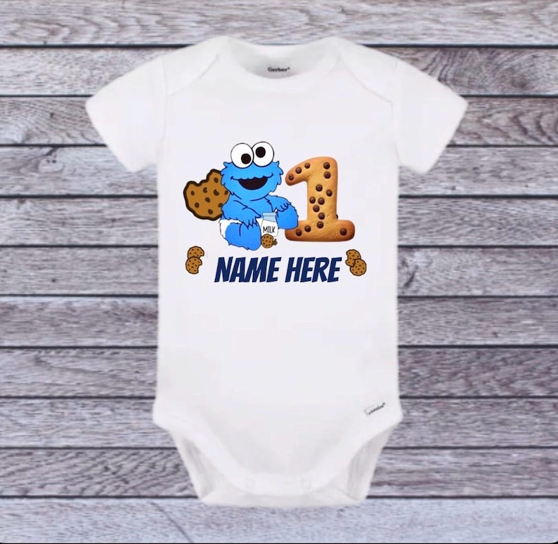 Pu&ograve; includere: Body da bambino bianco con un disegno blu di Cookie Monster che tiene in mano un cartone di latte e un grande numero 1 fatto di biscotti. Il testo "NAME HERE" &egrave; stampato in blu sotto il disegno. Sono presenti anche diverse grafiche di biscotti.