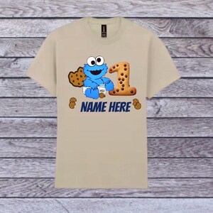 Pu&ograve; includere: T-shirt beige con un personaggio blu dei cartoni animati che tiene in mano un cartone di latte e un biscotto, con un grande numero 1 fatto di biscotti. Il testo "NAME HERE" &egrave; stampato in blu sotto. Piccole grafiche di biscotti sparse.