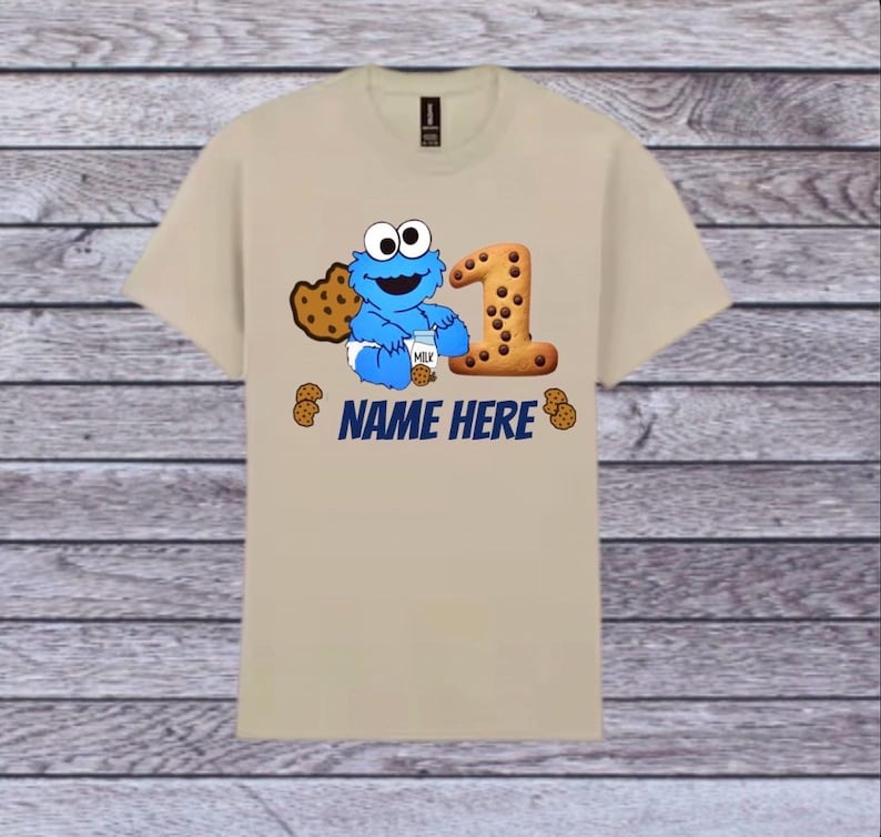 Pu&ograve; includere: T-shirt beige con un personaggio blu dei cartoni animati che tiene in mano un cartone di latte e un biscotto, con un grande numero 1 fatto di biscotti. Il testo "NAME HERE" &egrave; stampato in blu sotto. Piccole grafiche di biscotti sparse.