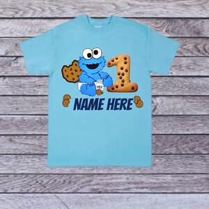 Pu&ograve; includere: Una maglietta azzurra con un design di Cookie Monster. Il design include un grande numero "1" fatto di biscotti, un biscotto e la scritta "NAME HERE". Cookie Monster tiene in mano un cartone di latte e un biscotto.