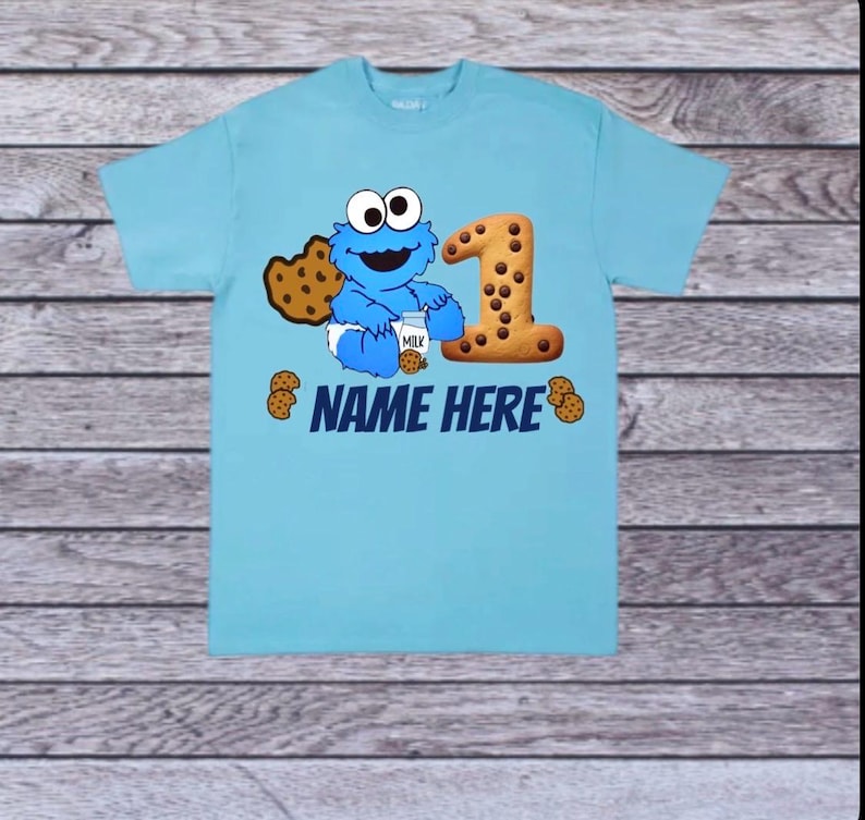 Pu&ograve; includere: Una maglietta azzurra con un design di Cookie Monster. Il design include un grande numero "1" fatto di biscotti, un biscotto e la scritta "NAME HERE". Cookie Monster tiene in mano un cartone di latte e un biscotto.
