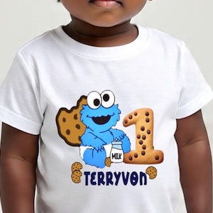 Pu&ograve; includere: T-shirt bianca con un design blu di Cookie Monster, un biscotto e il numero 1. La maglietta ha anche il nome "TERRYVON" stampato in blu. Il design include un cartone di latte e diversi biscotti con gocce di cioccolato.