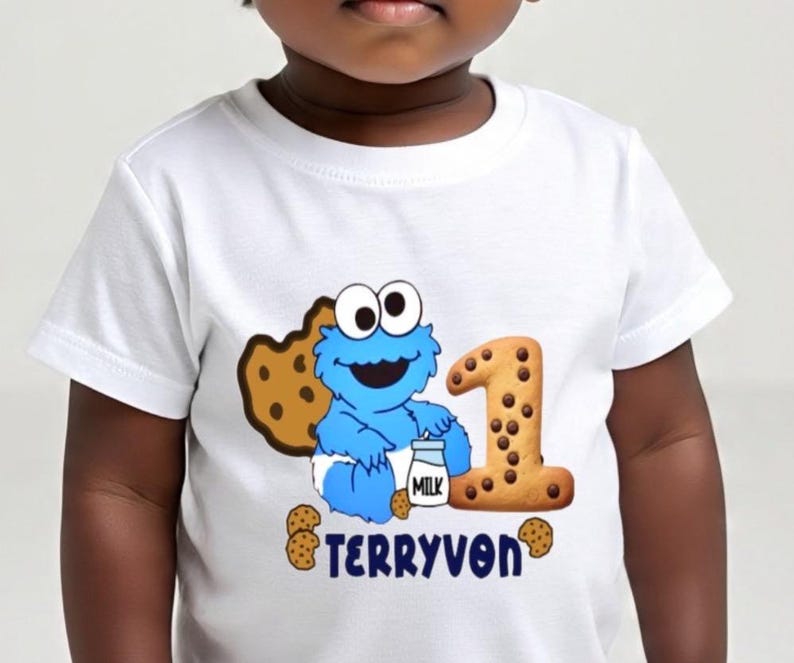 Pu&ograve; includere: T-shirt bianca con un design blu di Cookie Monster, un biscotto e il numero 1. La maglietta ha anche il nome "TERRYVON" stampato in blu. Il design include un cartone di latte e diversi biscotti con gocce di cioccolato.