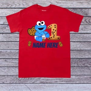 Pu&ograve; includere: T-shirt rosso con un design di Cookie Monster, un grande numero 1 fatto di biscotti e la scritta "NAME HERE" in blu. Il design &egrave; probabilmente per un primo compleanno. La maglietta &egrave; in materiale morbido.