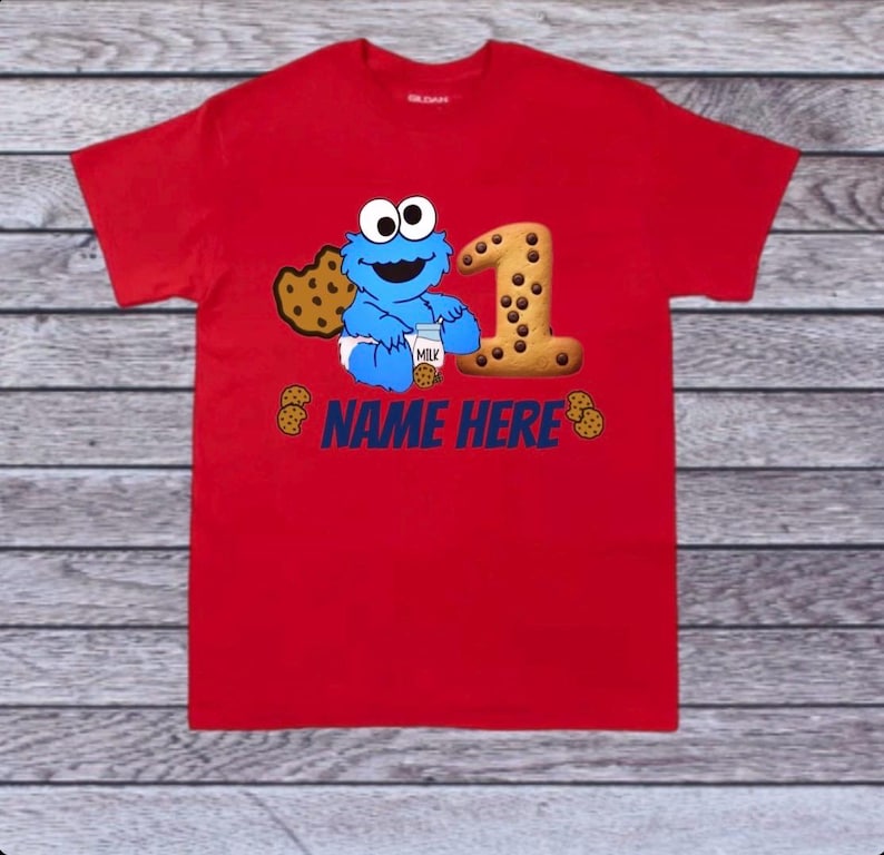 Pu&ograve; includere: T-shirt rosso con un design di Cookie Monster, un grande numero 1 fatto di biscotti e la scritta "NAME HERE" in blu. Il design &egrave; probabilmente per un primo compleanno. La maglietta &egrave; in materiale morbido.