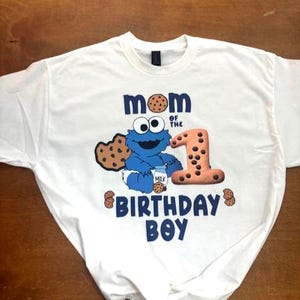 Pu&ograve; includere: T-shirt bianco con un disegno blu di Cookie Monster che tiene in mano un cartone di latte. Il testo sulla maglietta recita "MOM OF THE 1 BIRTHDAY BOY" in blu. Il numero 1 &egrave; disegnato per assomigliare a un biscotto.